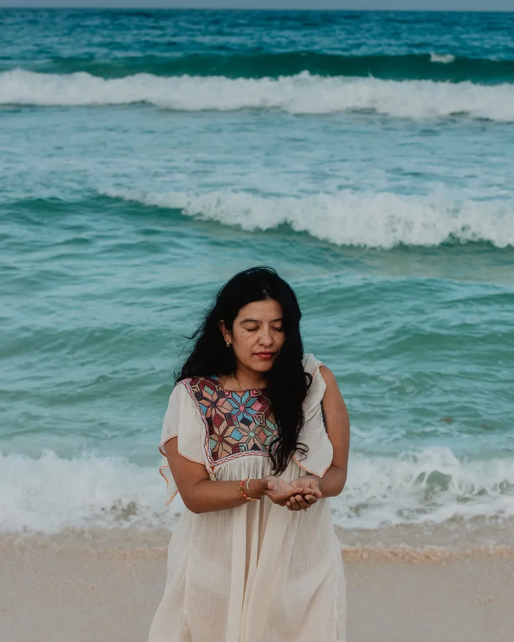 Retrato de Blanca Coutiño de pie frente al mar, vestido de lino con bordado maya, sosteniendo cristales en sus manos ahuecadas
